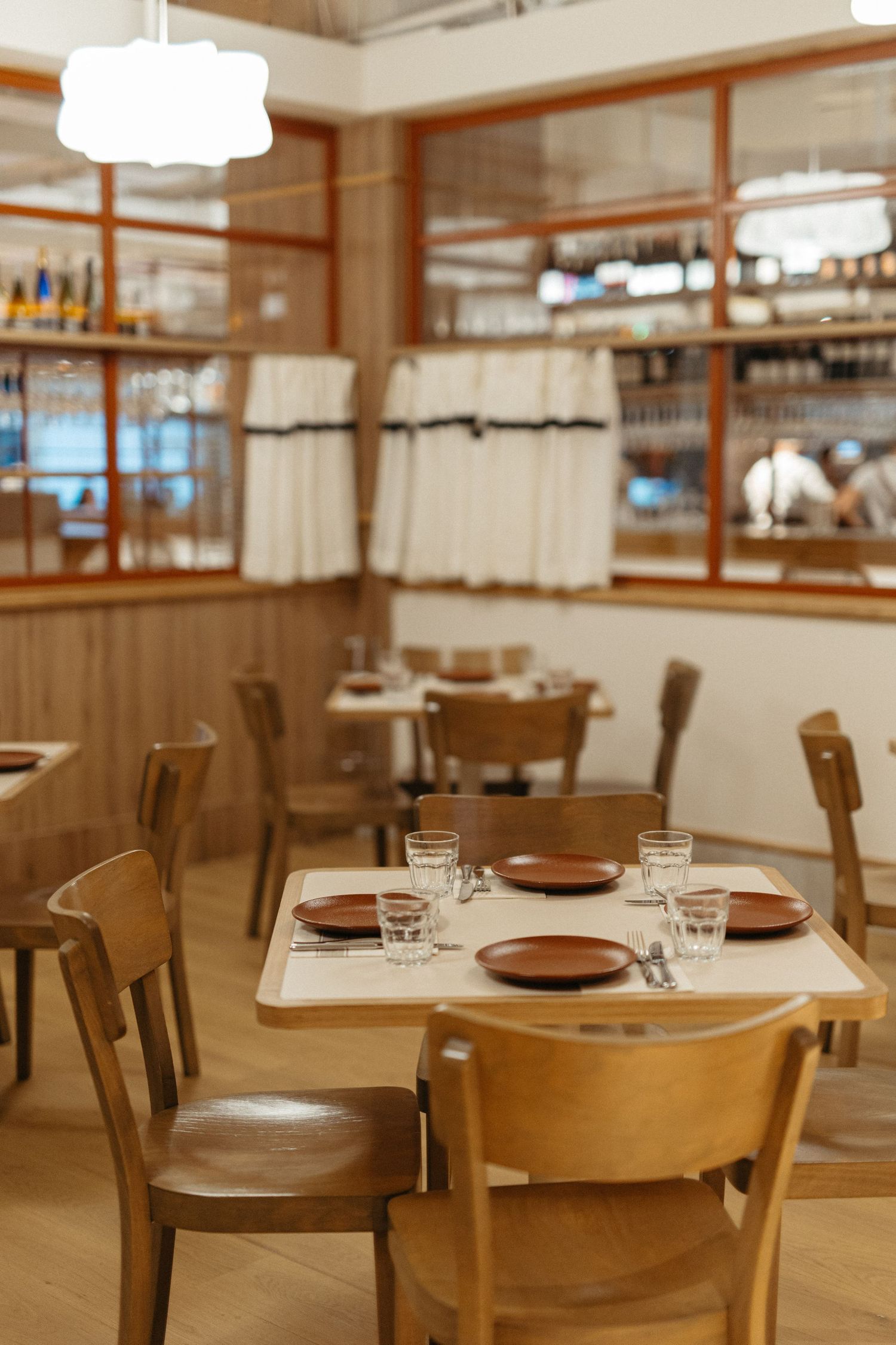 Cozy restaurant interior with wooden chairs, set tables featuring brown plates and glasses, wine shelves, and curtained windows.