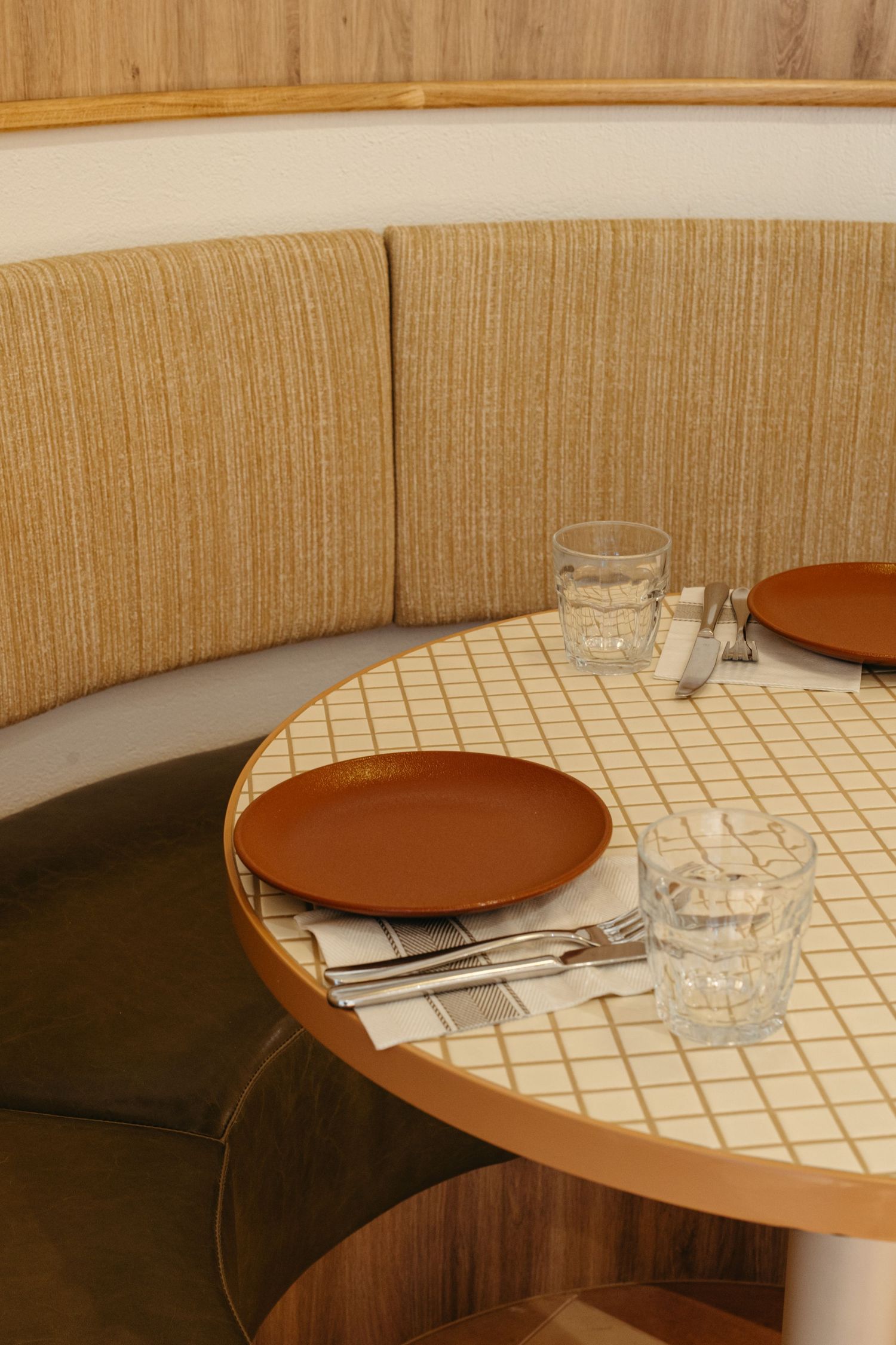 Curved banquette booth with beige upholstery beside a round tiled table set with terracotta plates and glass tumblers.