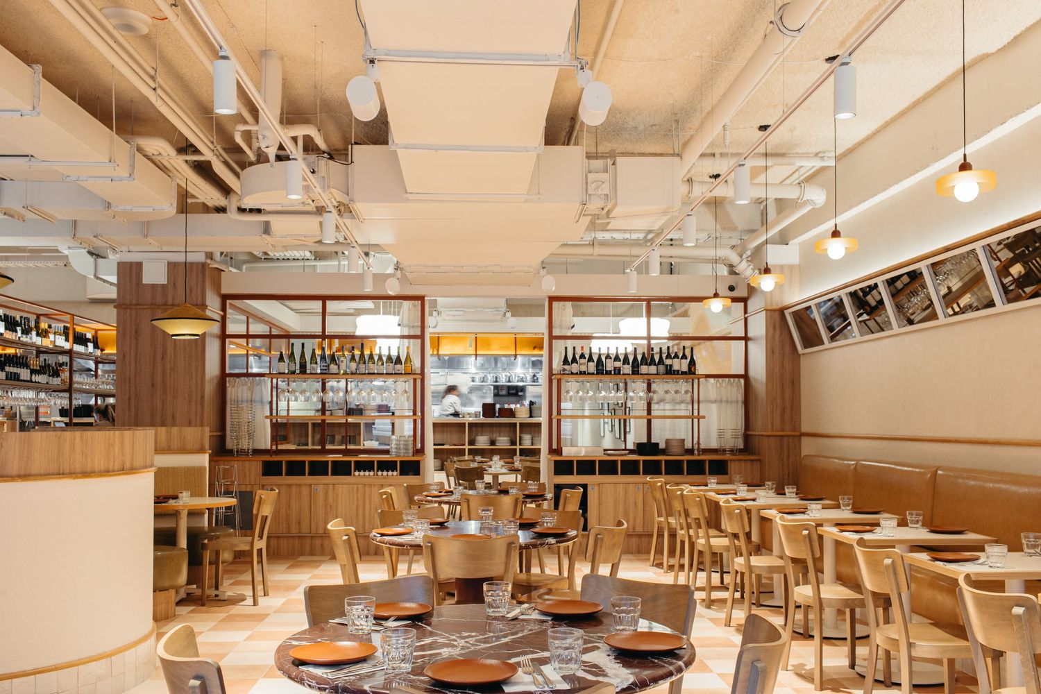 Modern restaurant interior with wooden chairs, round tables, open kitchen, and warm pendant lighting.