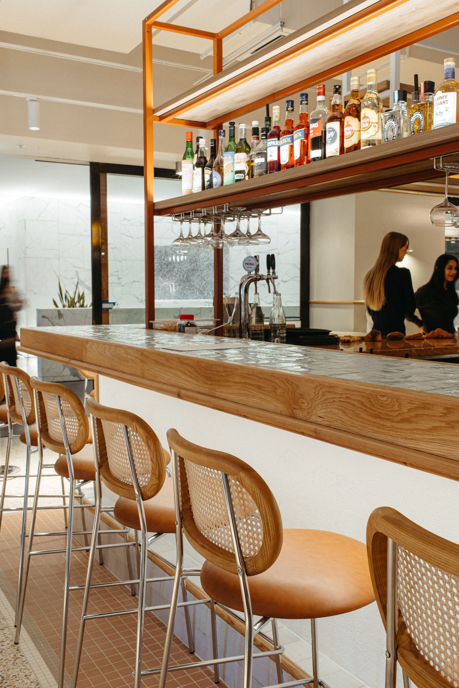 Modern bar with rattan stools, wooden counter, overhead liquor shelf, and bartenders working in bright interior.