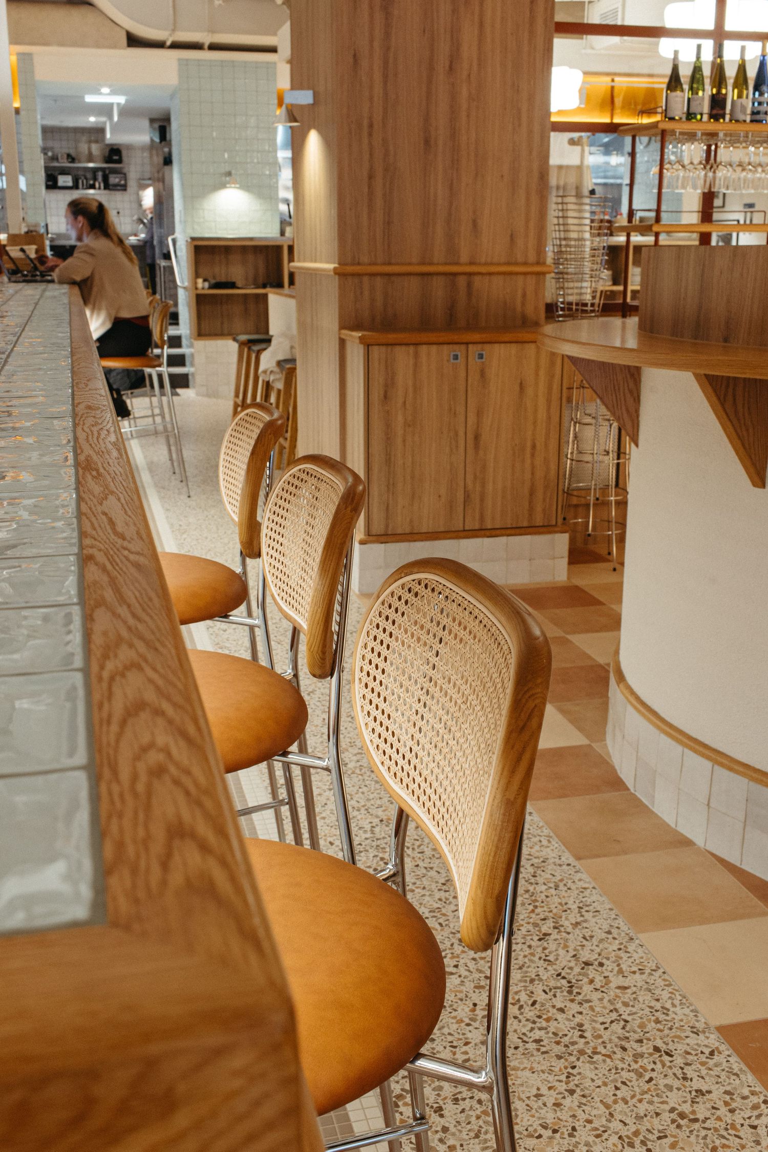 Modern bar with rattan counter stools, warm wood accents, terrazzo floors, and a woman dining in the background.