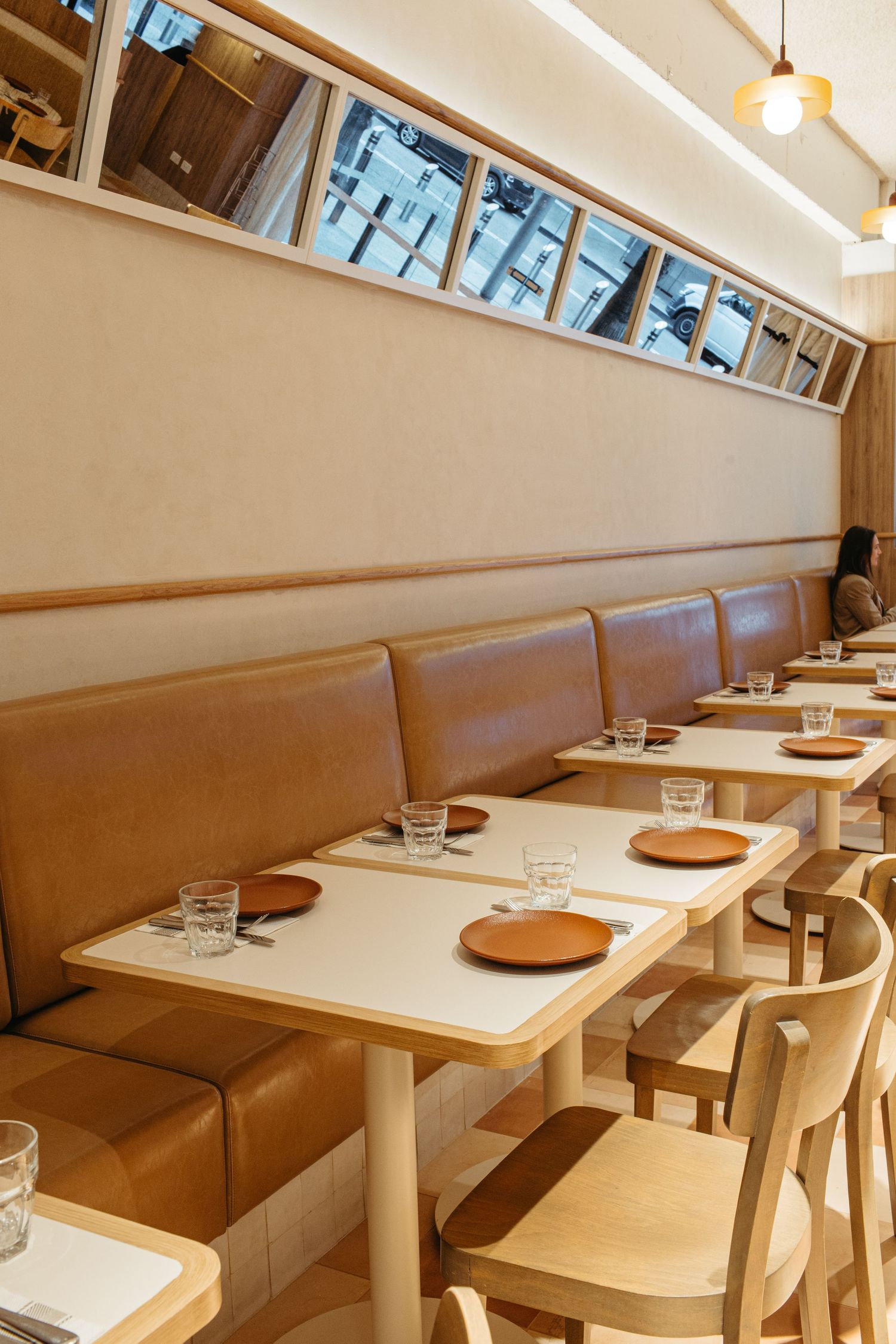 Modern restaurant interior with tan leather banquette seating, light wood chairs, and white tables set with glasses and plates.