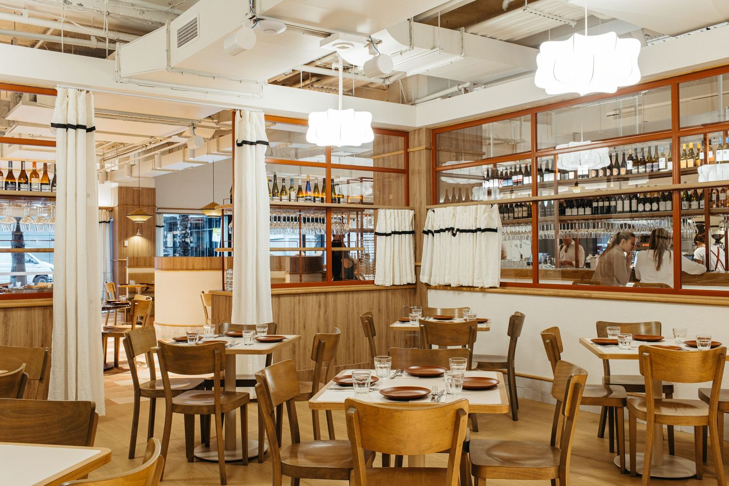 Bright modern restaurant interior with wooden tables, chairs, white curtains, and a wine display shelf backdrop.