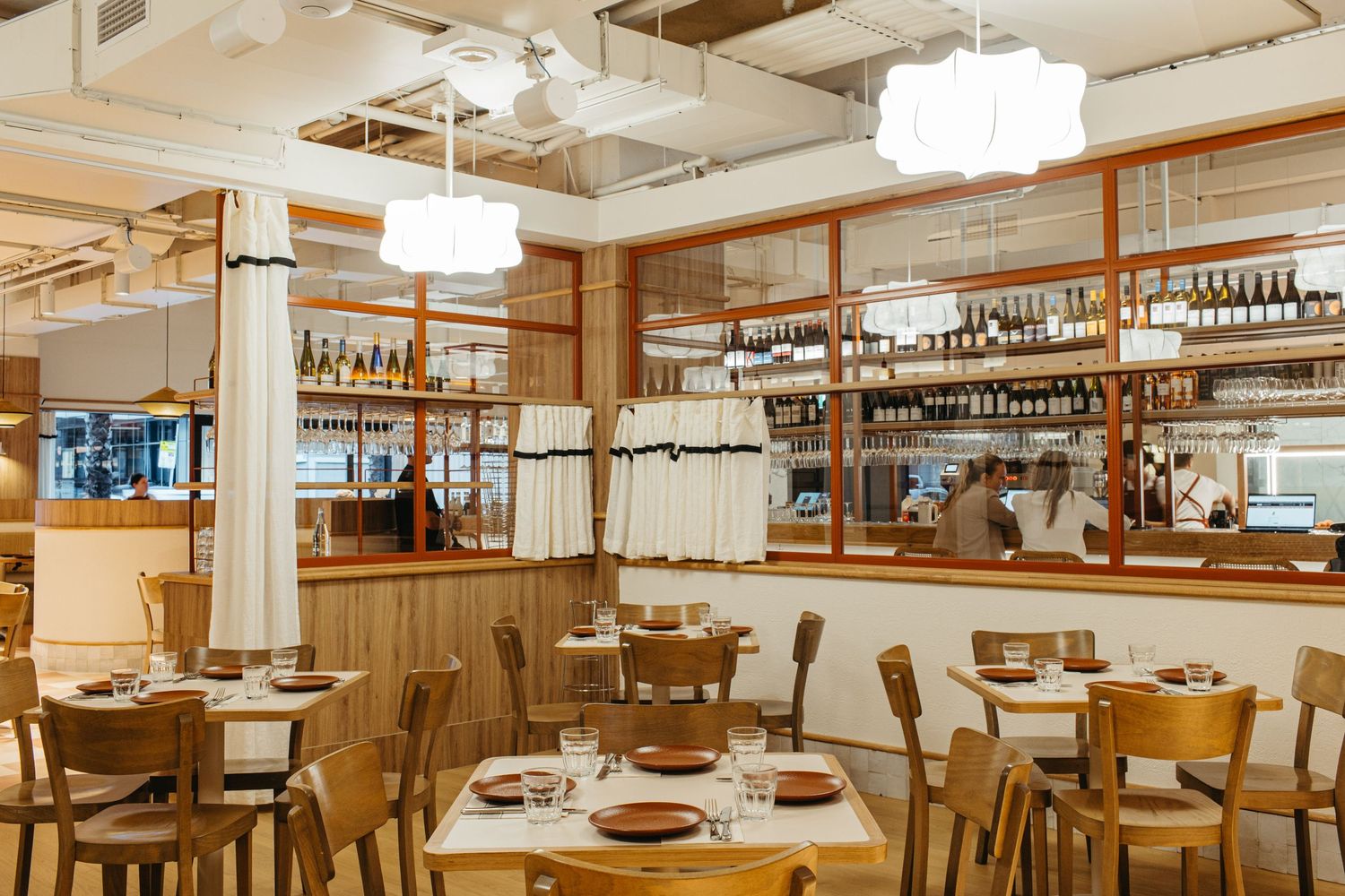 Modern restaurant interior with wooden furniture, pendant lights, open kitchen, and wine bottle display shelves.