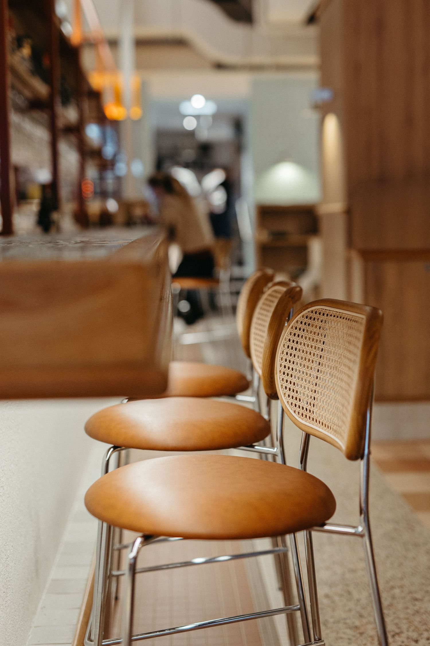 Modern bar stools with tan leather seats and wicker backs on chrome frames lined up along a wooden counter in a cafe.