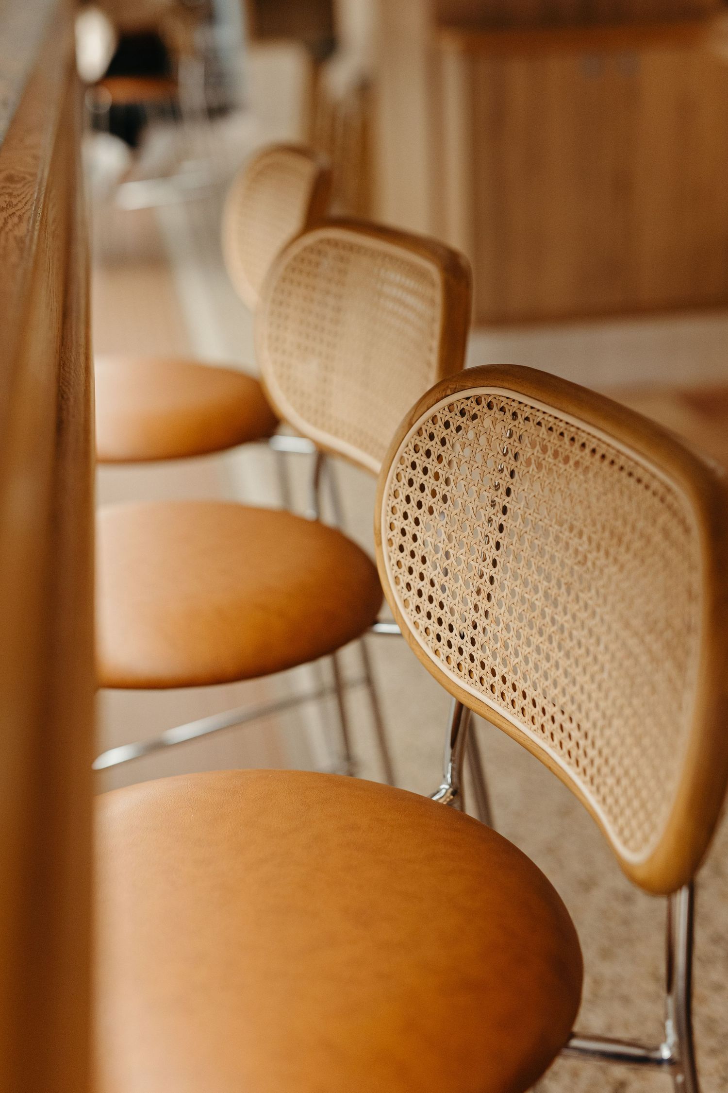 Row of elegant bar stools with tan leather seats and woven cane backs on chrome frames along a wooden counter.