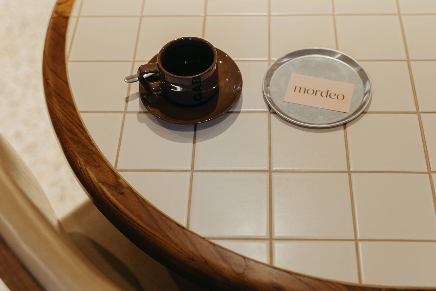 A small black espresso cup on a tiled table next to a round Mordeo branded coaster.