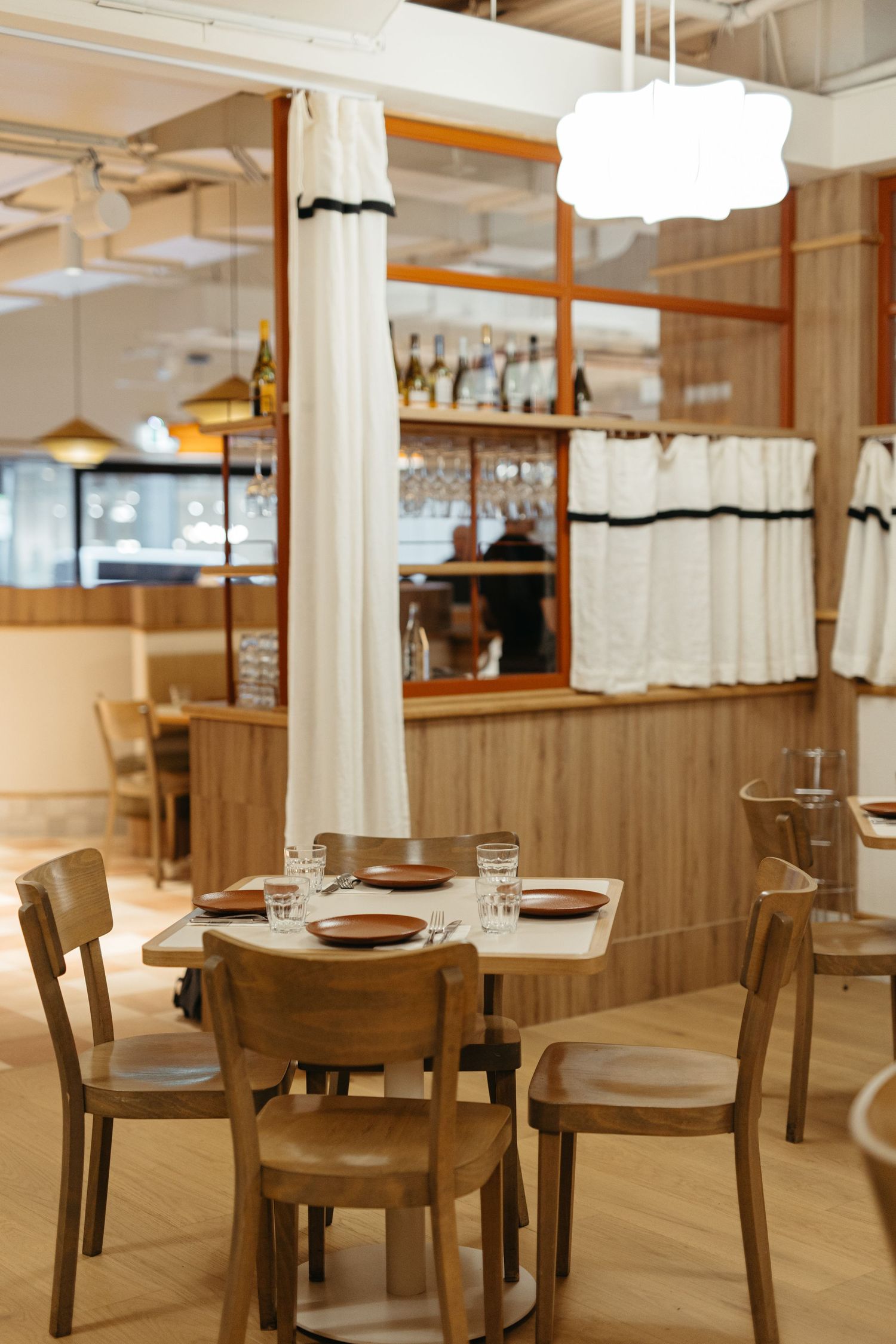 Cozy restaurant interior with wooden chairs, set tables, white curtains, and a bar area in warm neutral tones.