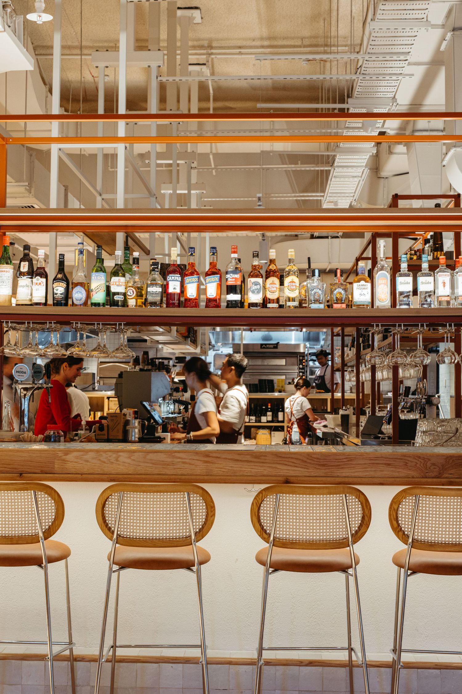 Modern restaurant bar with rattan stools, shelves of liquor bottles, and bartenders working behind a wooden counter.