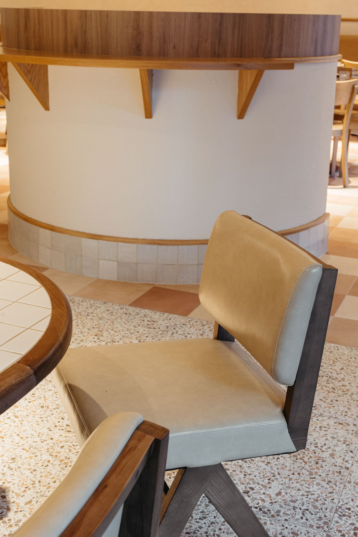 Modern restaurant interior with beige upholstered chair, wooden frame, terrazzo floor, and curved white pillar accent wall.