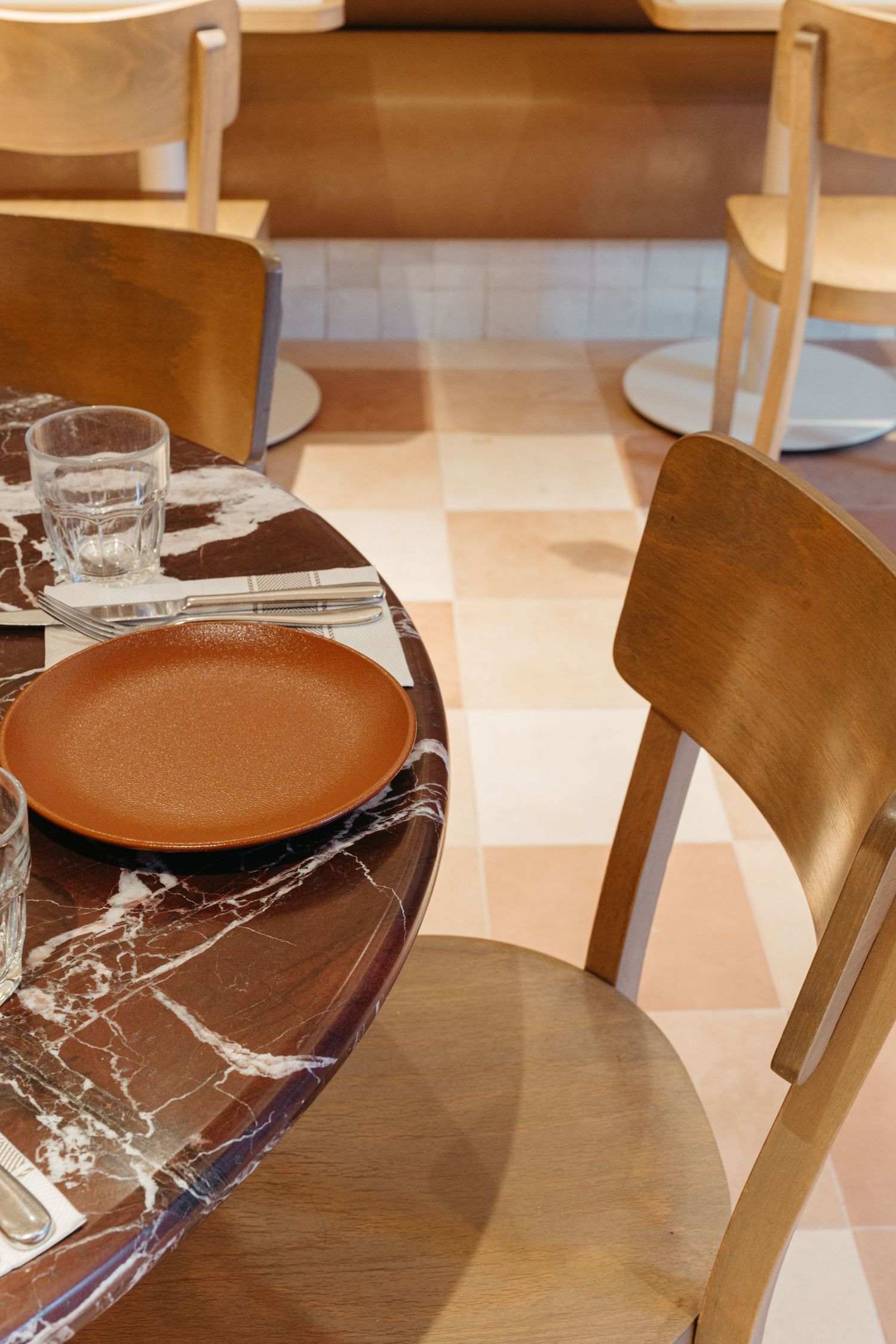 Elegant restaurant table setting with marble top, terracotta plate, glass, and wooden chairs on checkered floor.