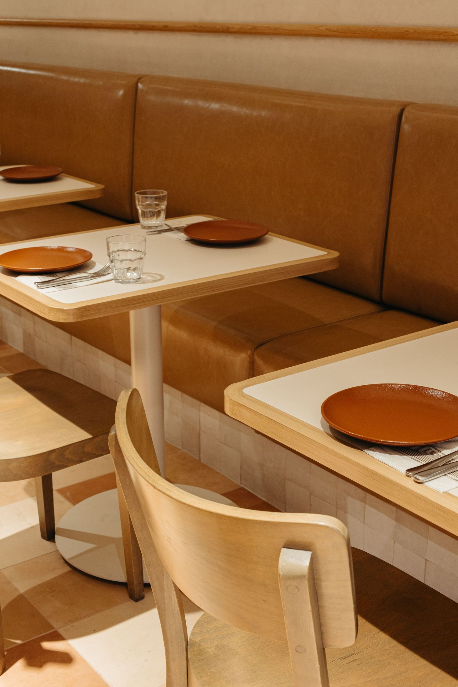 Modern restaurant booth seating with tan leather banquette, wooden chairs, and terracotta plates on white tables.