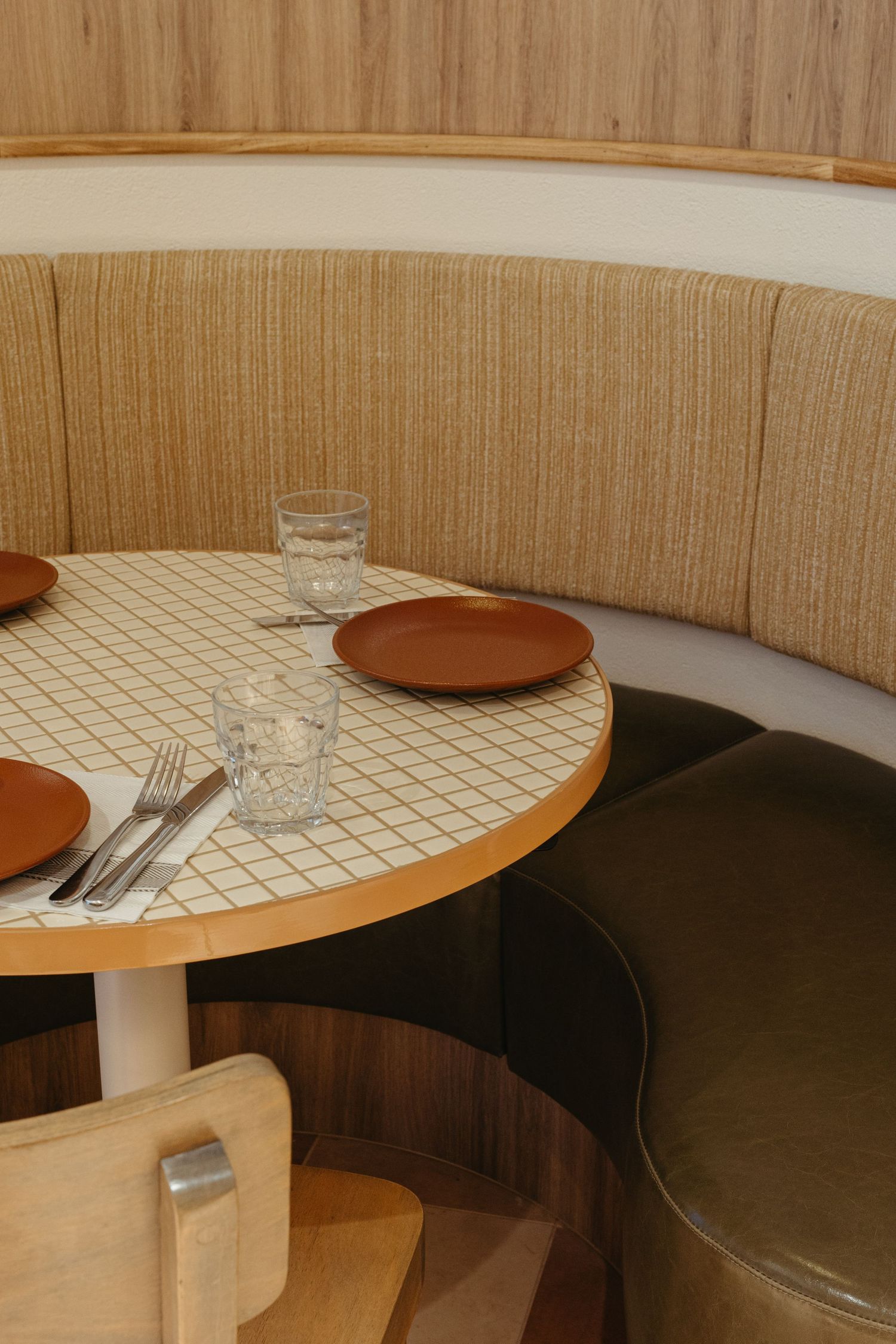 Cozy restaurant booth with curved beige upholstered seating, round tile-top table, terracotta plates, and crystal glasses.