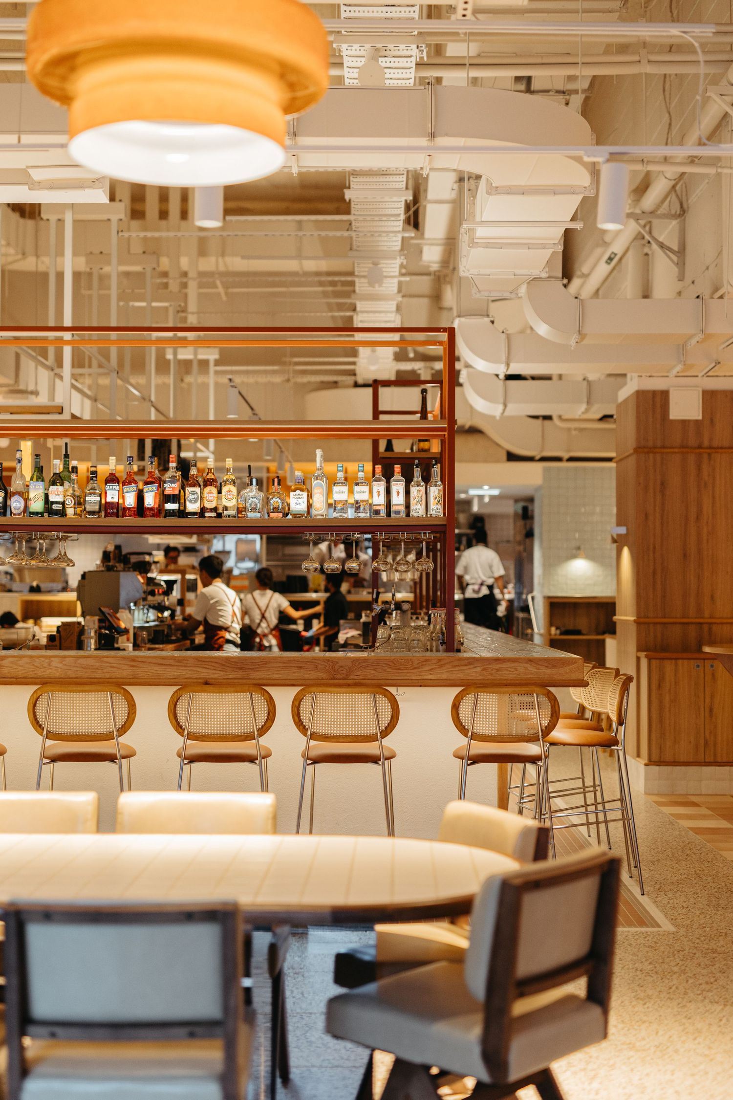 Modern restaurant bar with rattan stools, wooden counter, bottle display, and warm pendant lighting in an open-plan dining space.