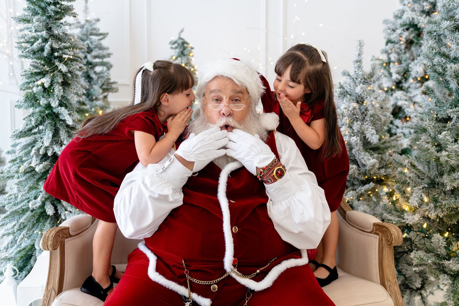 Two girls in red dresses whisper to Santa Claus seated in a chair surrounded by decorated Christmas trees.