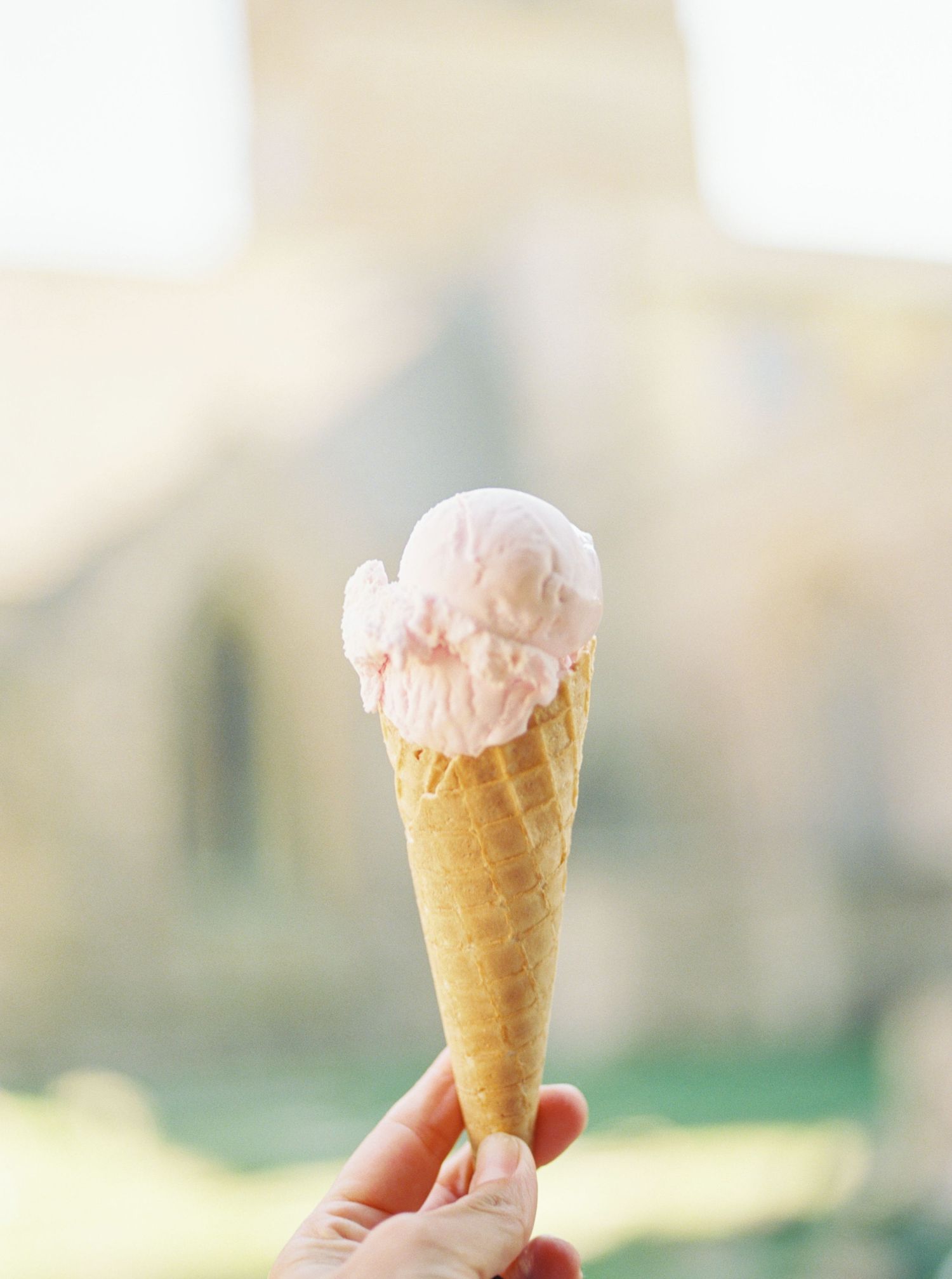 A hand holding a waffle cone with two scoops of pink strawberry ice cream against a soft blurred outdoor background.