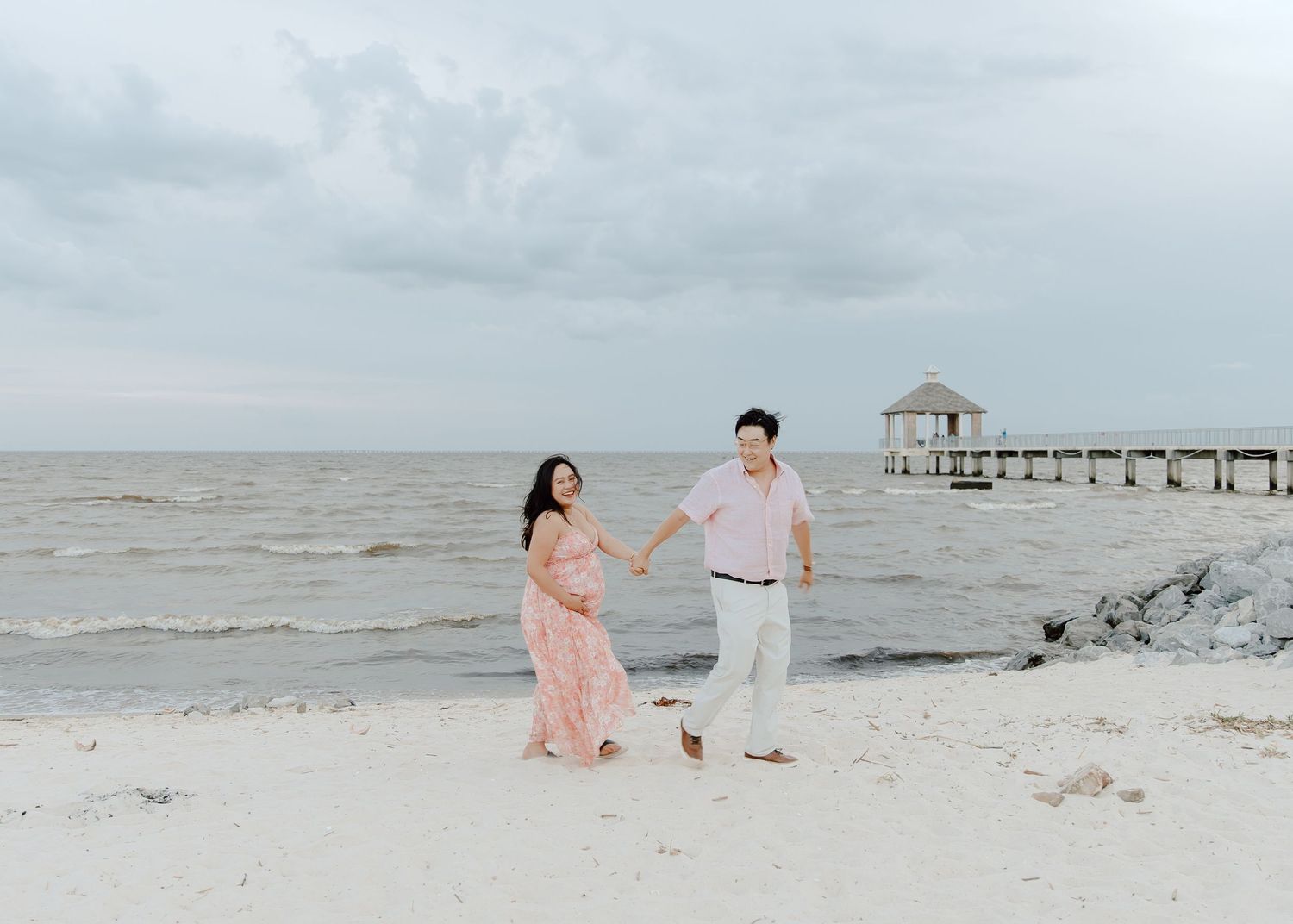 Couple holding hands on a white sand beach, woman in pink flowy dress, man in light pink shirt, with a pier in the background.