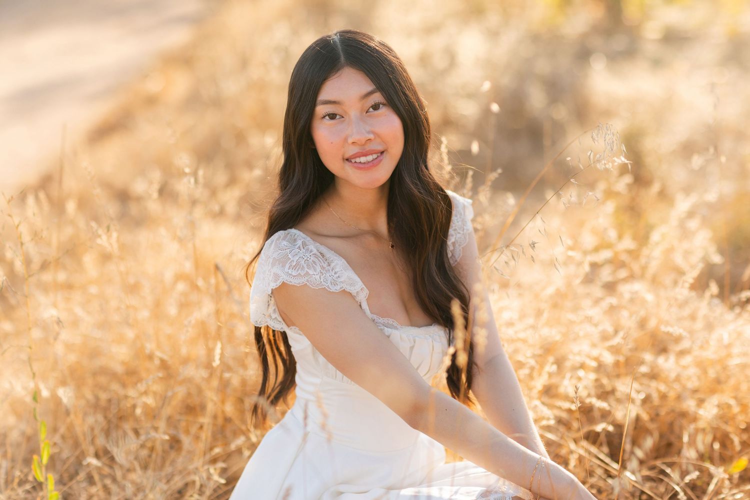 Young Asian woman in white lace dress sitting in golden sunlit field, smiling gracefully at camera.