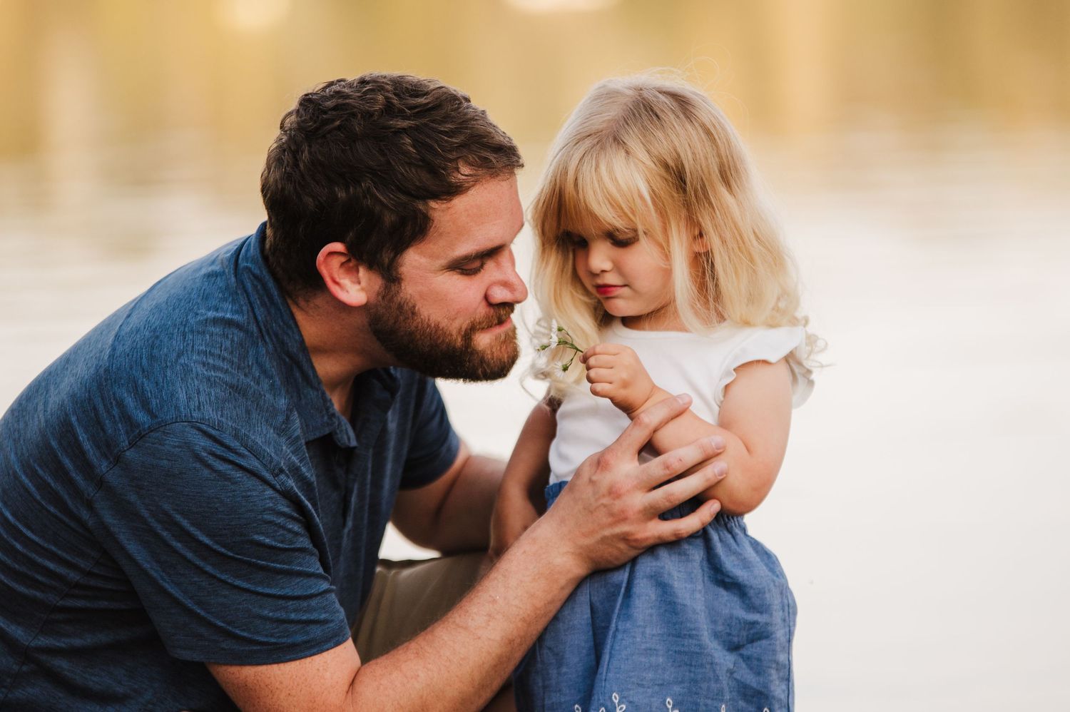 A father tenderly kisses his young blonde daughter outdoors near a lake in warm golden light.