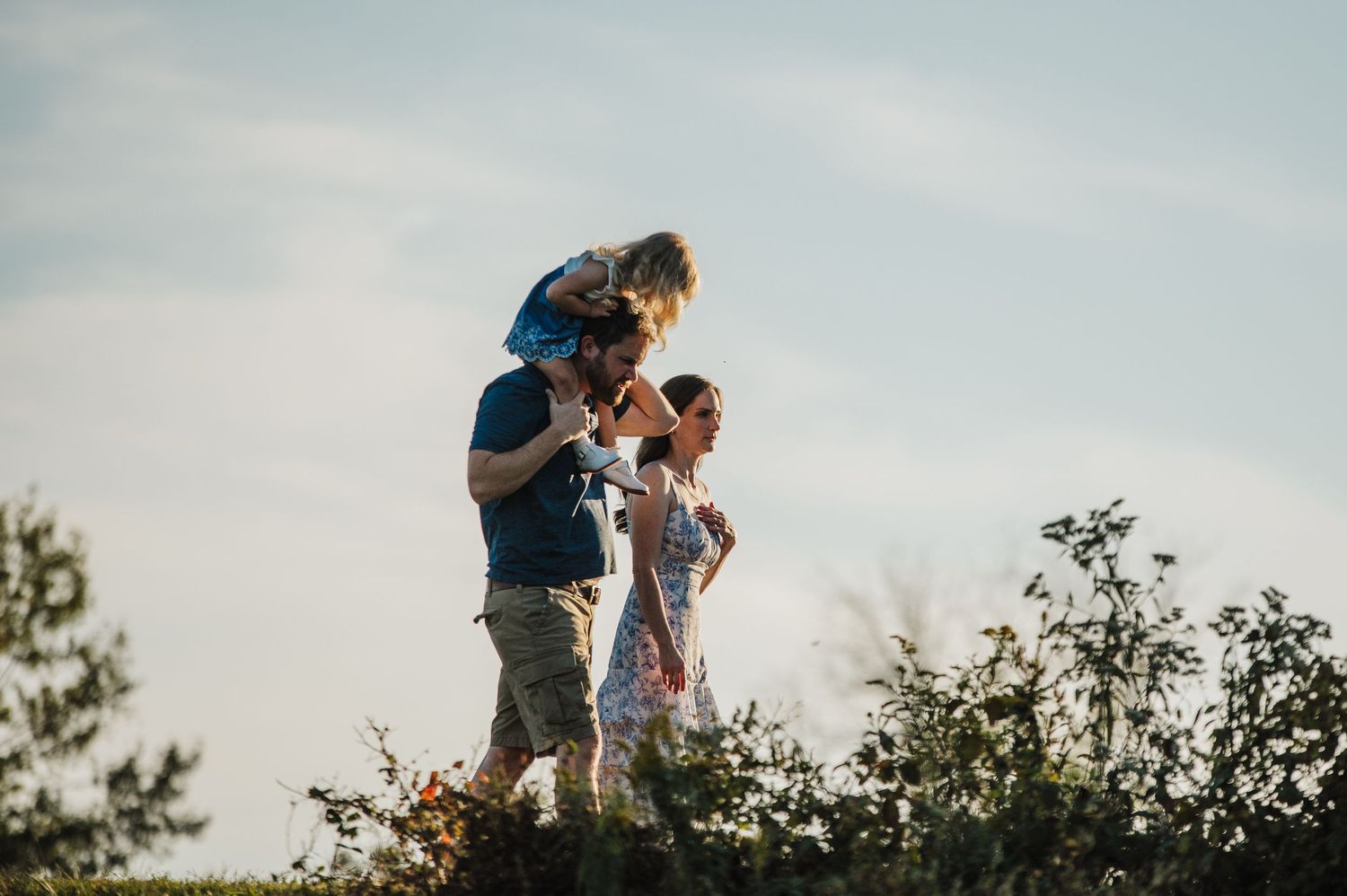 A family walks outdoors, with a child sitting on a man's shoulders beside a woman, surrounded by greenery under a bright sky.
