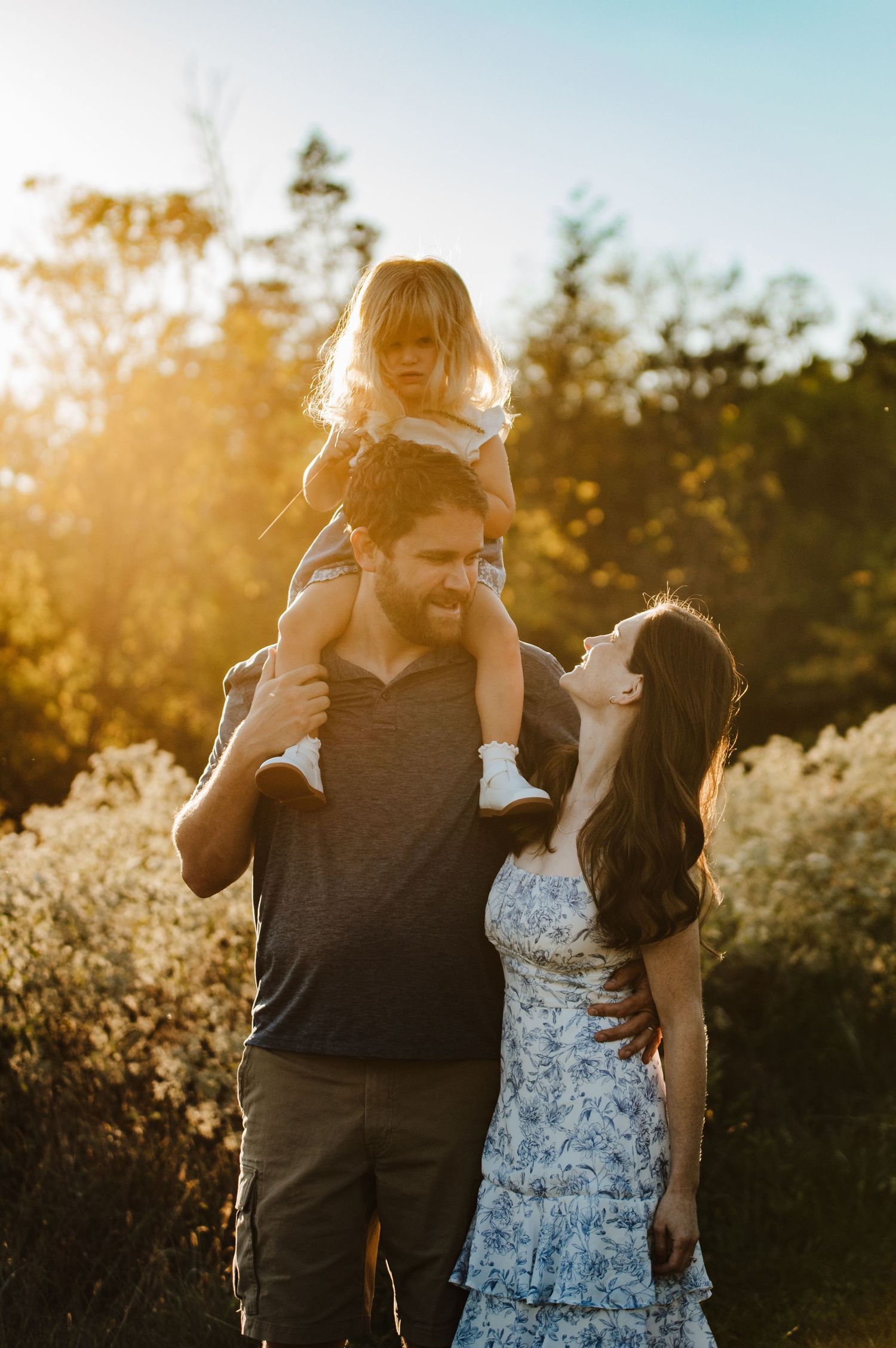 A joyful family at golden hour, dad carrying toddler on shoulders while mom laughs in a floral dress outdoors.
