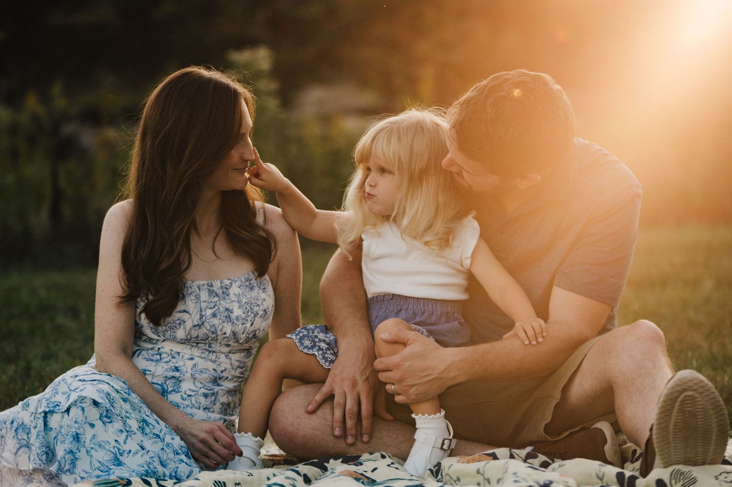 A happy family sitting on a blanket in sunlit park, toddler touching mom's face while dad looks on lovingly.