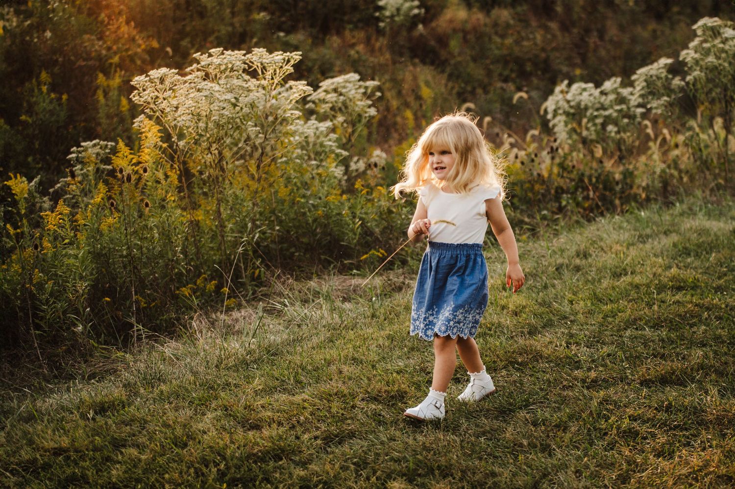 A young blonde girl in a blue skirt walks through a lush green meadow with wildflowers at golden hour.