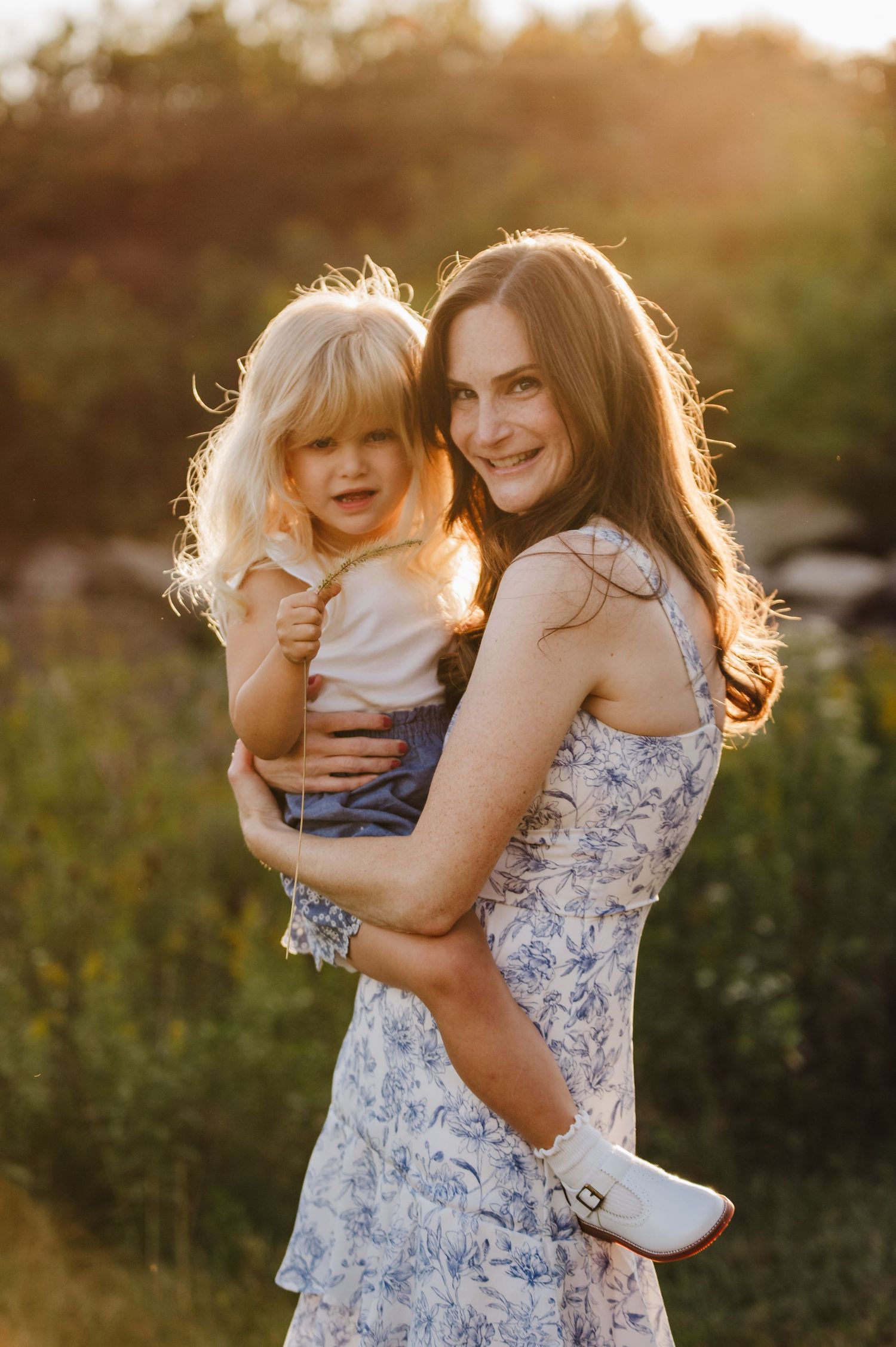 A smiling brunette woman in a floral dress holds a blonde toddler in a sunlit outdoor setting.