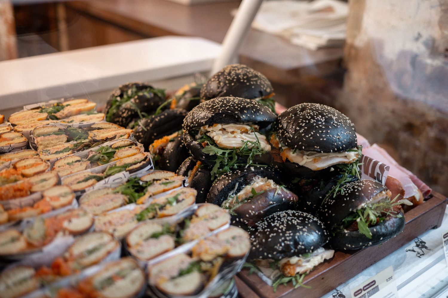 A display of black sesame seed burgers and assorted sandwiches piled on a wooden tray at a food market.