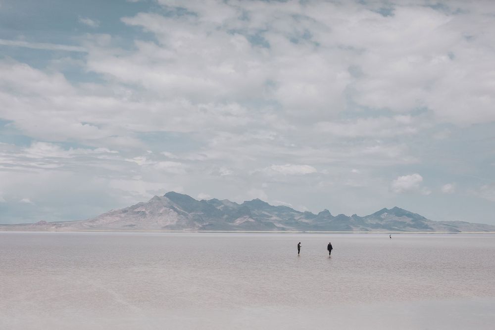 Two distant figures walking across vast white salt flats with mountains and cloudy sky in background.