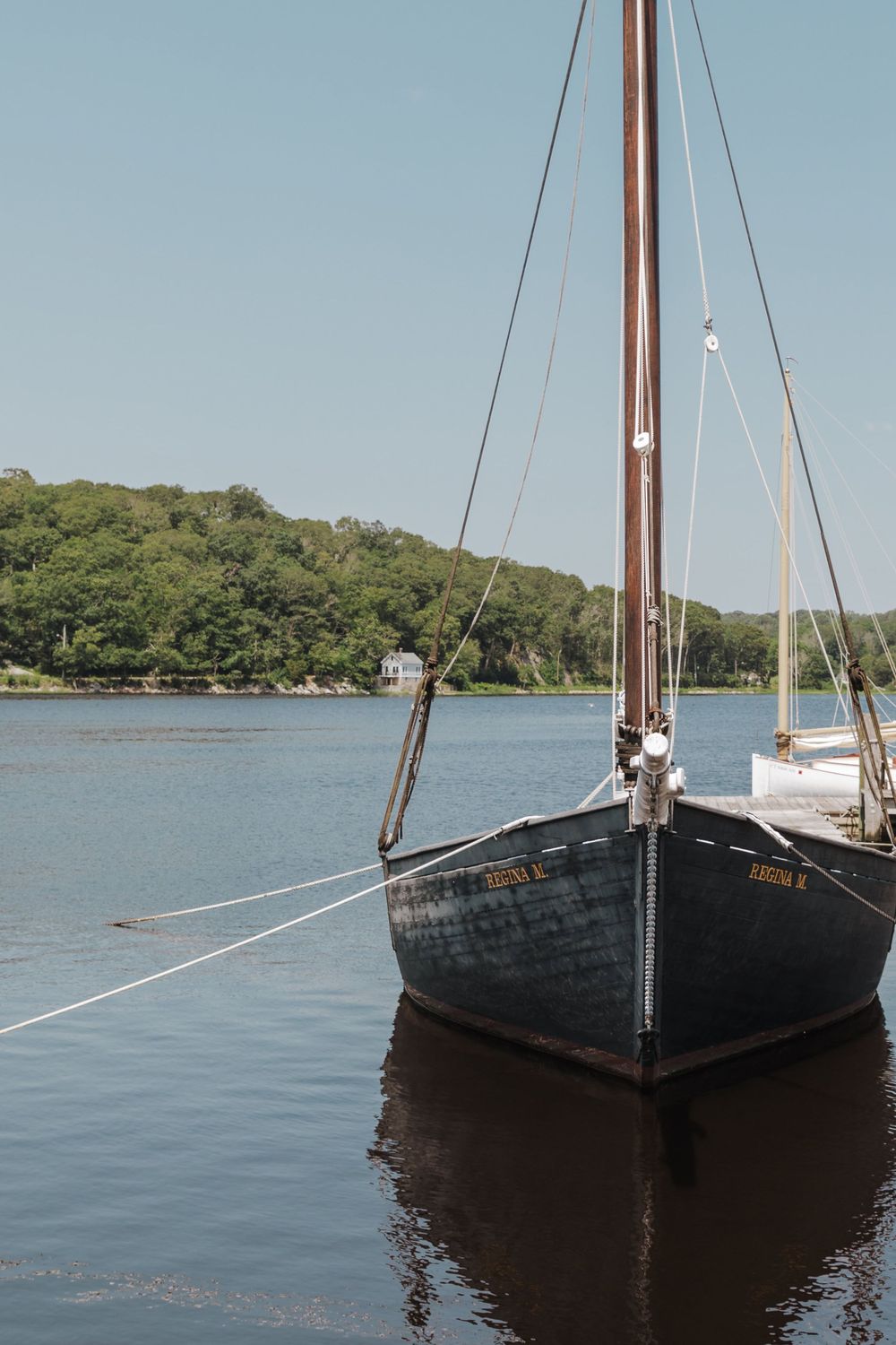 A classic wooden sailboat with dark hull sits moored on calm waters against a tree-lined shore on a serene day.