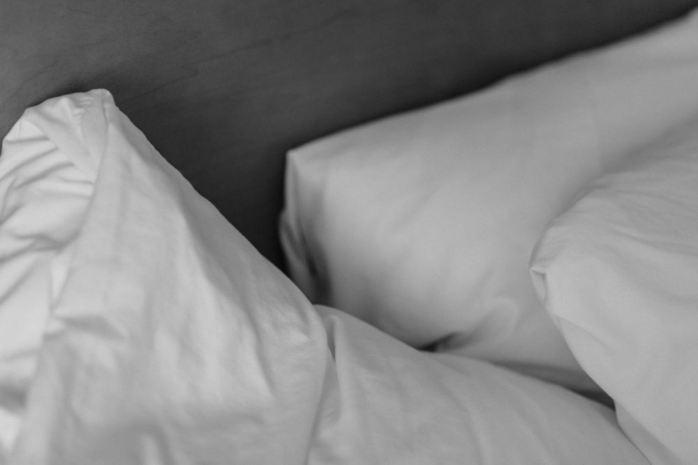 Close-up black and white photograph of wrinkled white bedsheets and pillows showing texture and shadows.