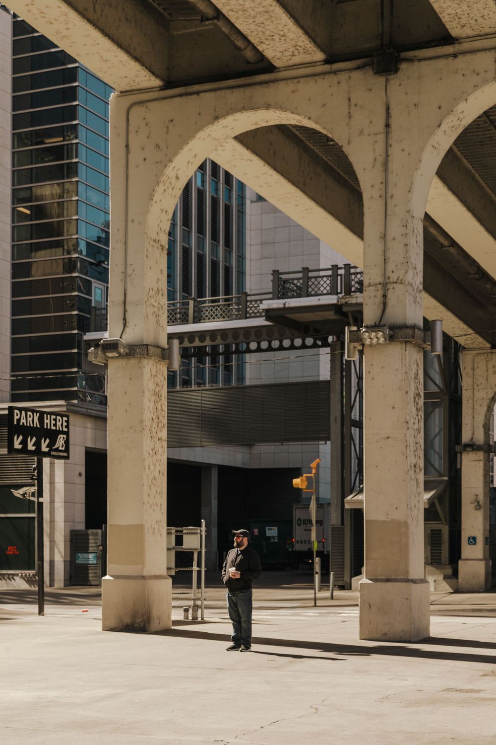 Person standing beneath concrete arches of urban architecture with modern buildings and blue glass windows in background.