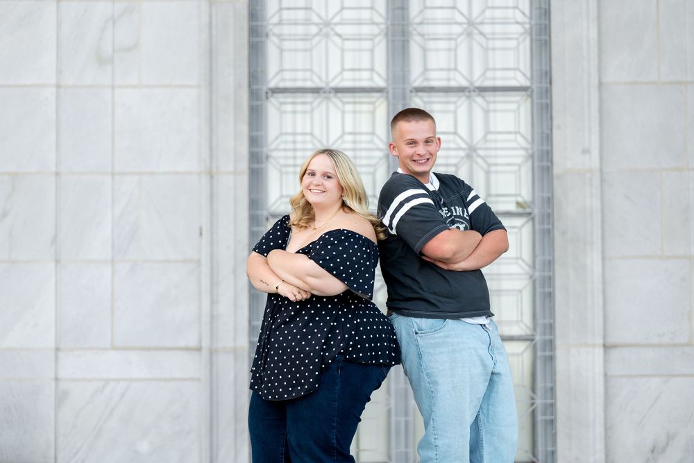 Couple posing back-to-back with arms crossed in front of ornate marble building doors.