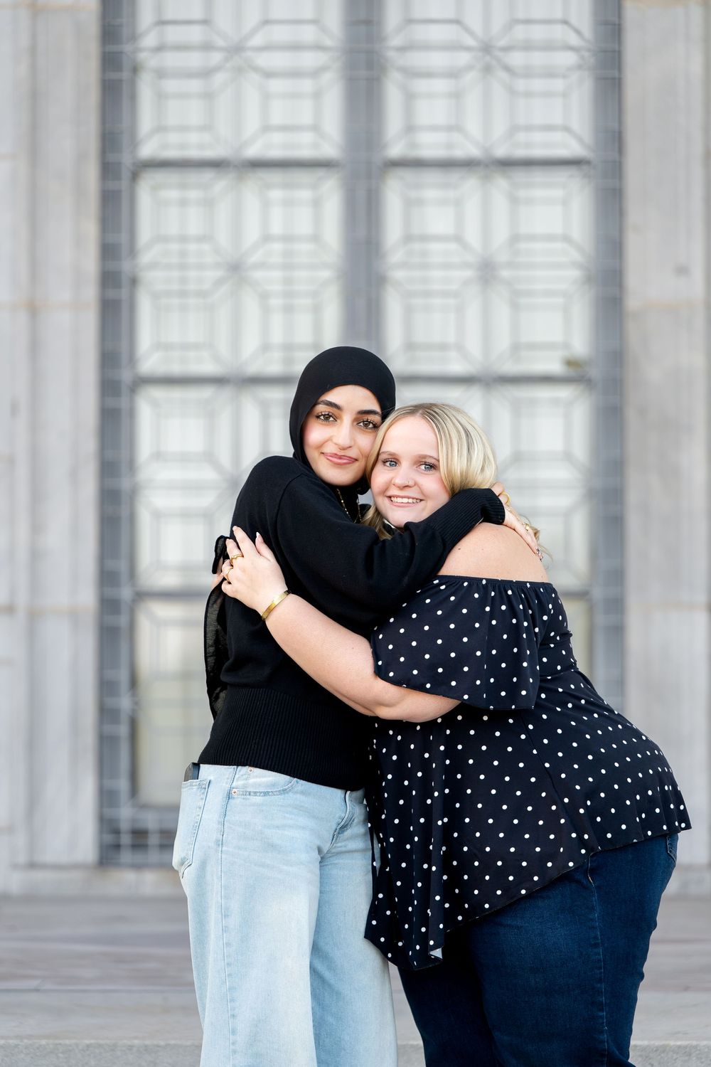 Two young women share a warm hug outdoors, one wearing a black hijab and the other a polka dot top.