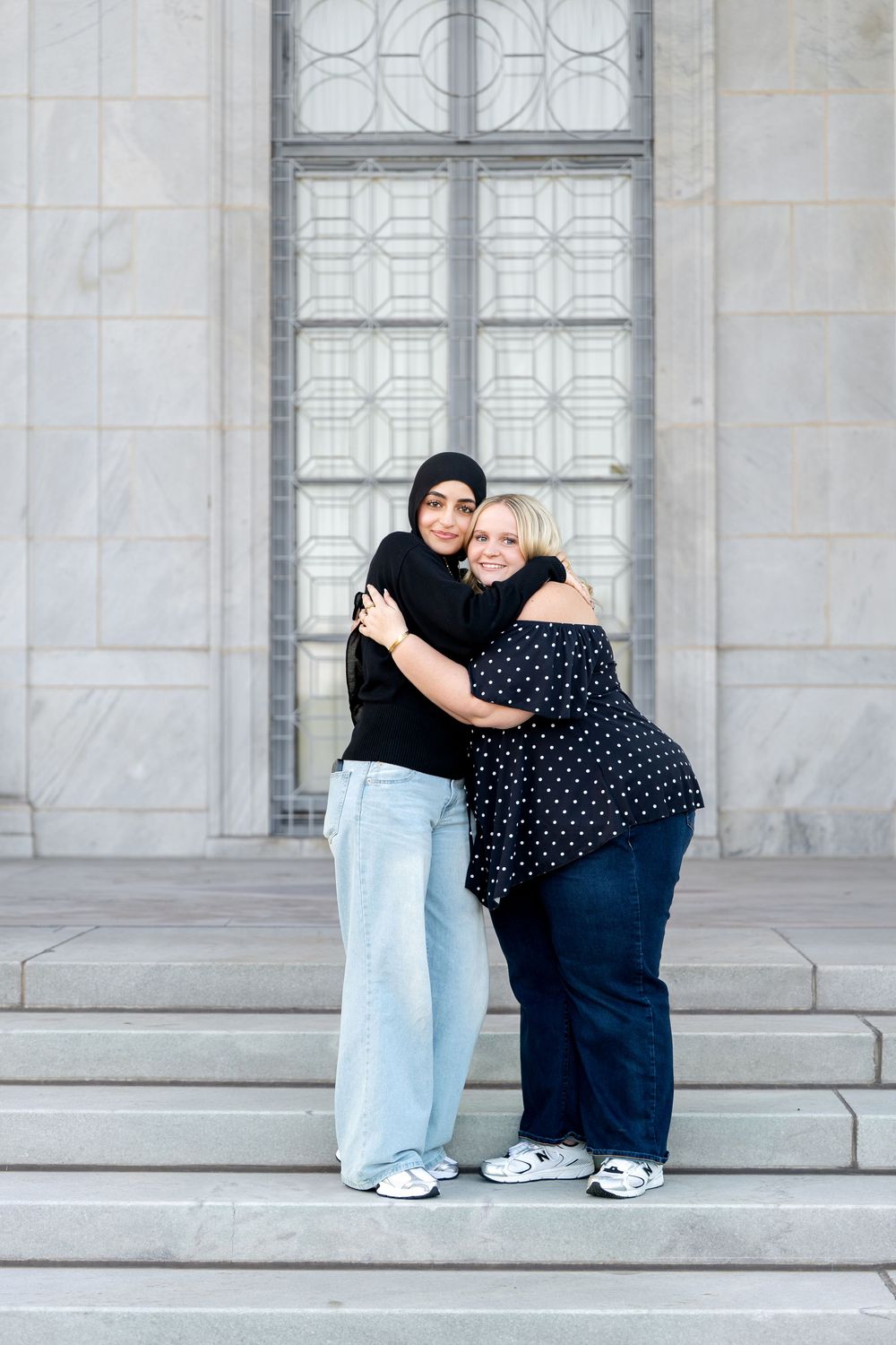 Two women hugging and smiling on marble steps, one wearing a black hijab and jeans, the other in a polka dot blouse.