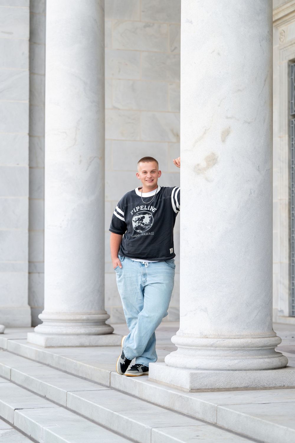 Smiling teenage boy in black jersey and jeans leaning against marble columns on steps.