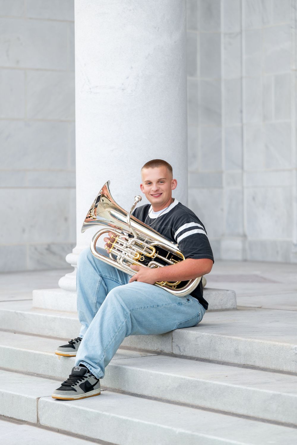 Smiling teen boy in black jersey sitting on marble steps holding a euphonium instrument.