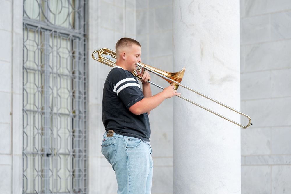 Young man in black t-shirt playing trombone outdoors near white marble columns in daylight.