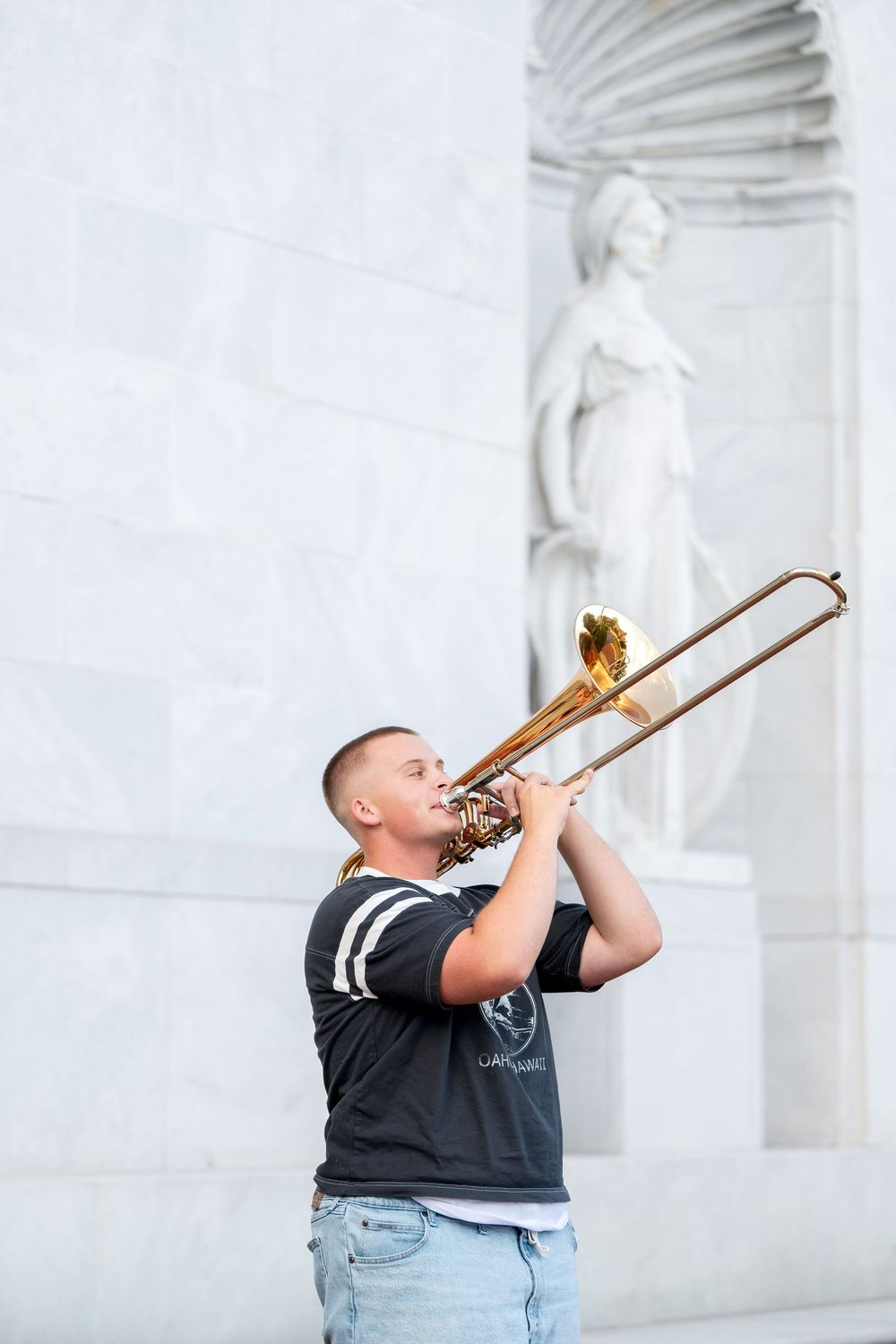 Young musician plays trombone outdoors near classical marble statue architecture.
