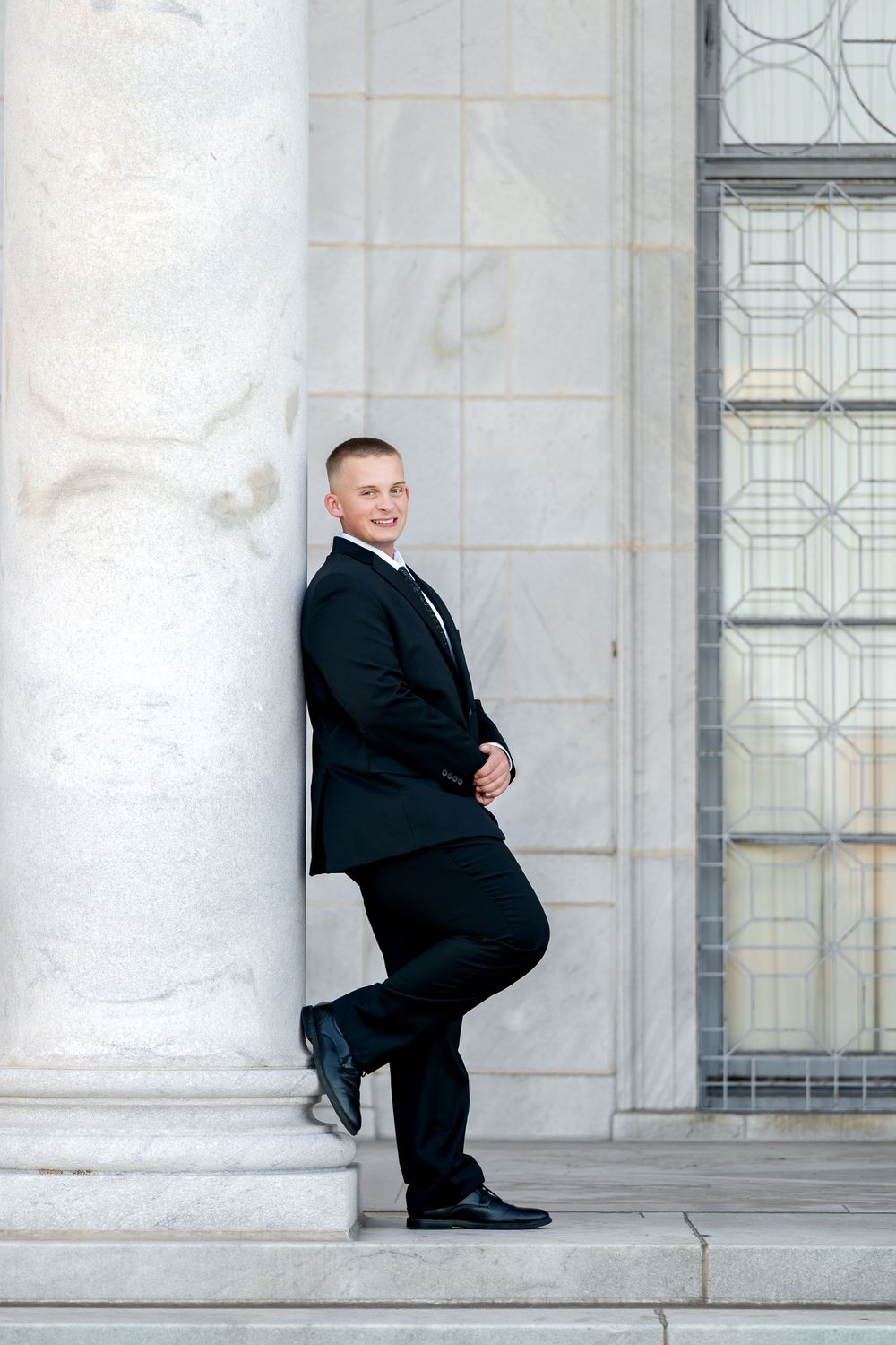Smiling young man in black suit leaning against marble column outside elegant building with decorative window.
