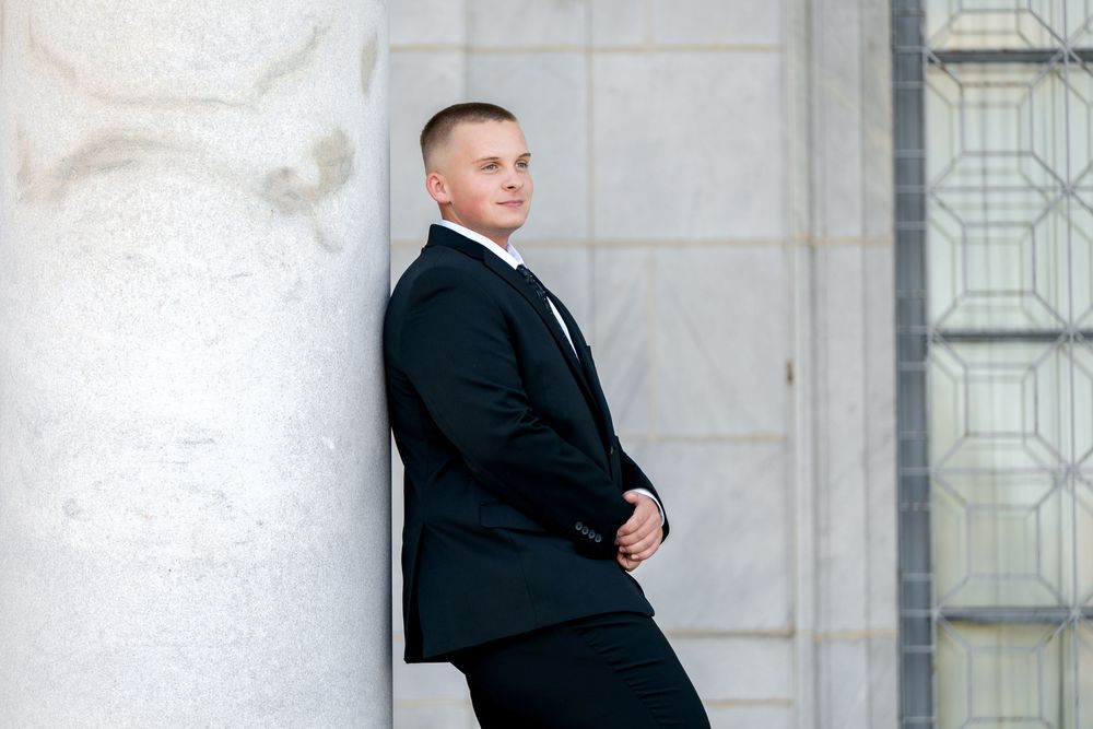 Young man in black suit leaning against marble column with a serious expression.
