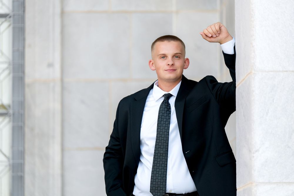 A confident young man in a black suit and tie poses with raised fist against marble columns.
