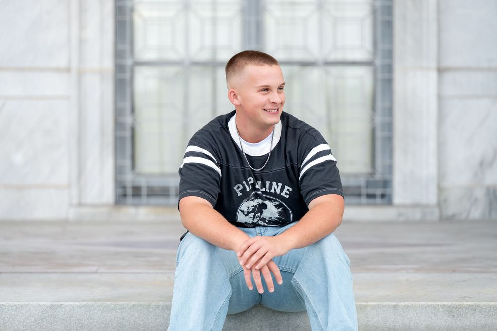 A young man in a Pipeline graphic tee and light jeans sits relaxed on marble steps smiling.