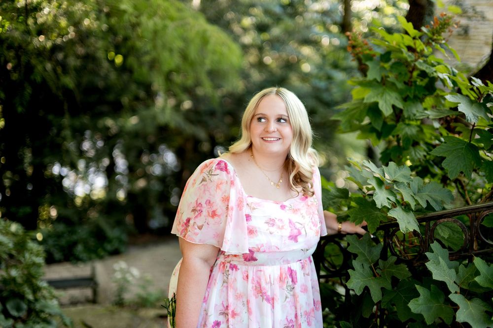 A smiling young blonde woman in a pink floral dress stands among green garden foliage.