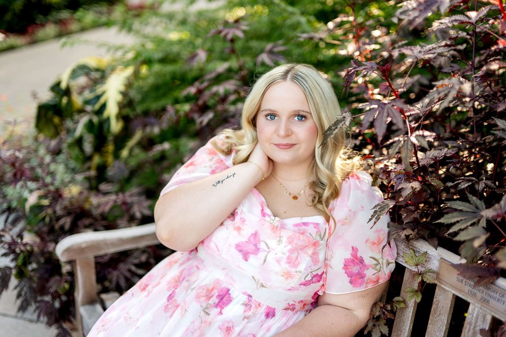 Young blonde woman in pink floral dress seated on wooden bench surrounded by lush green and purple garden foliage.