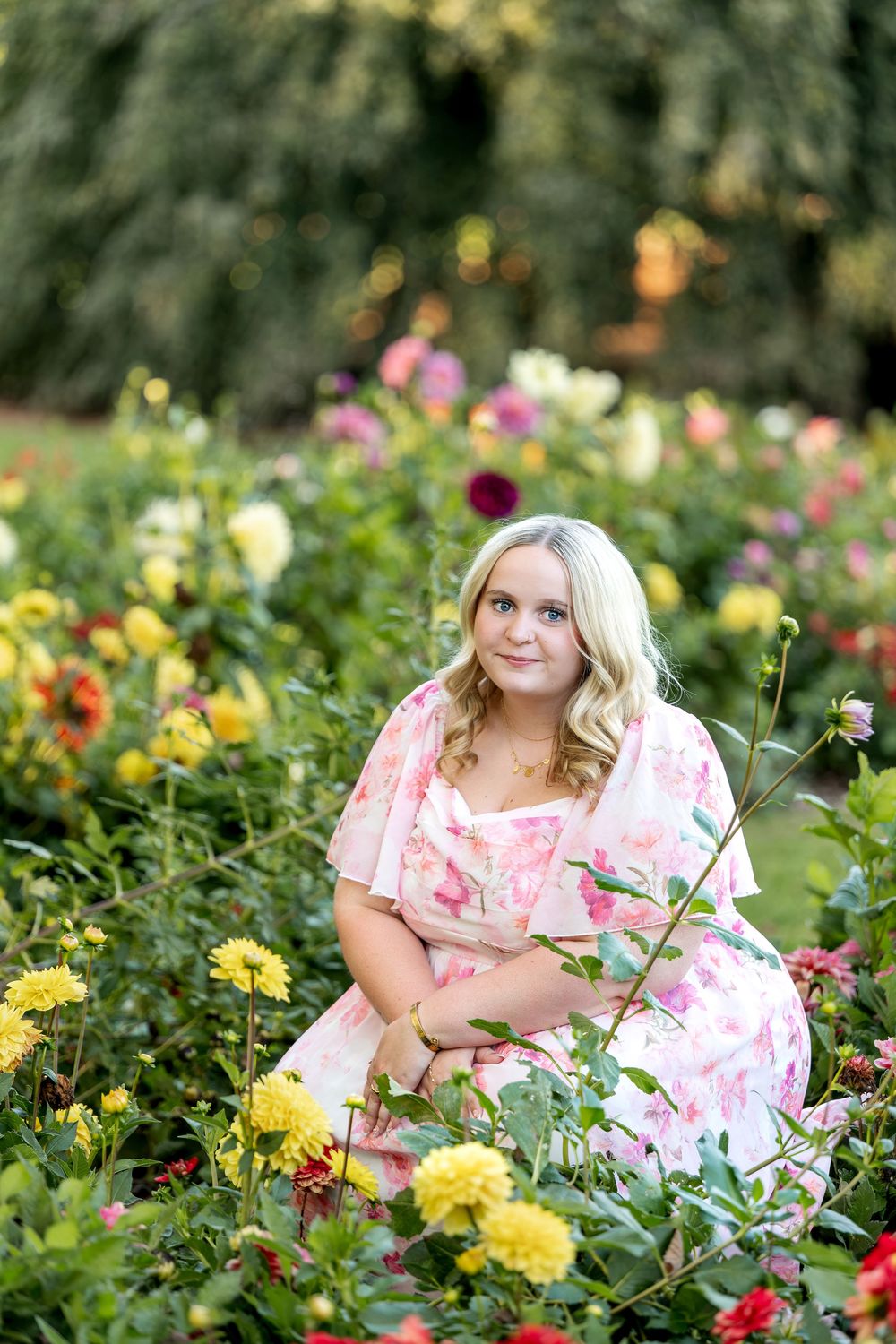 A young blonde woman in a pink floral dress kneels among colorful dahlia flowers in a garden.