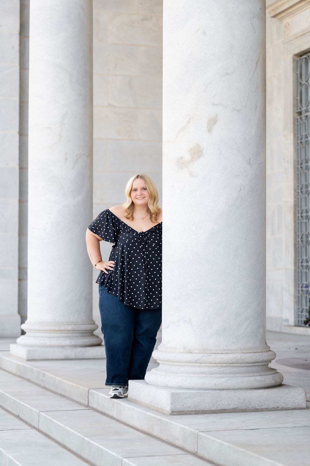 Blonde woman in black polka dot top and navy pants posing between marble columns on stone steps.