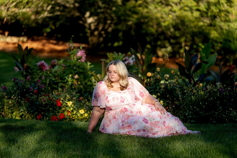 Young woman in pink floral dress sitting gracefully on green grass among colorful garden flowers.