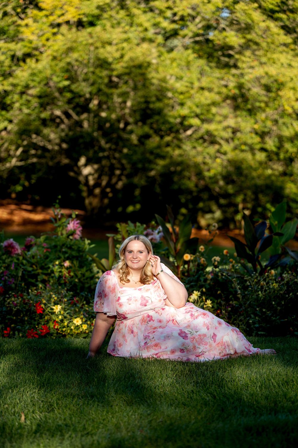 Young blonde woman in floral pink dress sits on grass in a lush garden.
